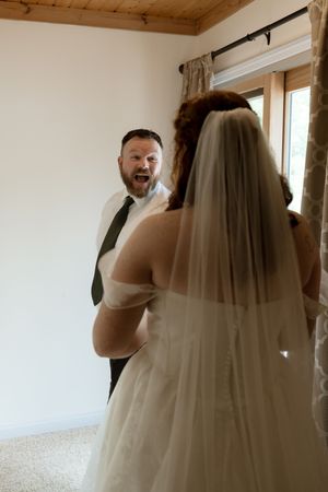 Groom reacts with joyful expression seeing bride in white wedding gown and veil during first look reveal.