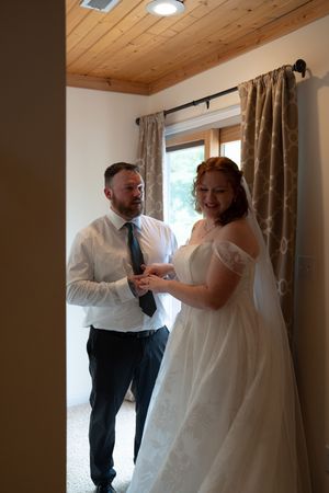 A groom places a wedding ring on his bride's finger as she smiles in her white off-shoulder gown and veil.