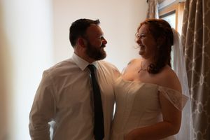 Bride in white off-shoulder dress and groom in white shirt and tie share a joyful glance by a sunlit window.