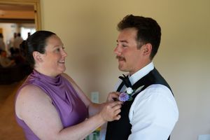 Woman in purple dress pinning boutonniere on groom in black vest and white shirt at wedding preparation.