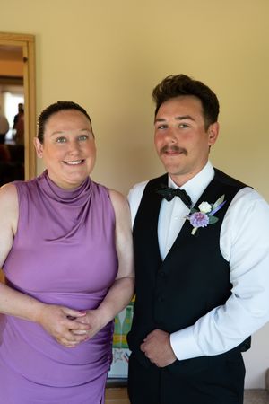 Woman in purple dress stands smiling beside groom in black vest with floral boutonniere indoors before wedding.
