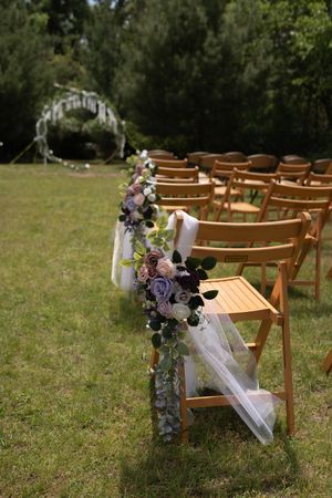 Elegant outdoor wedding aisle decorated with purple and mauve floral arrangements on wooden folding chairs.