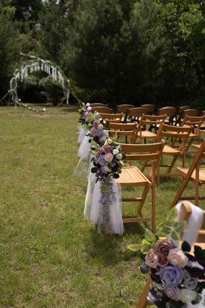 Elegant outdoor wedding ceremony aisle lined with wooden chairs adorned with purple and pink floral arrangements on green lawn.