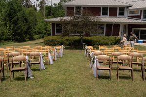 Outdoor wedding ceremony setup with wooden folding chairs tied with white ribbon bows on green lawn before red building.