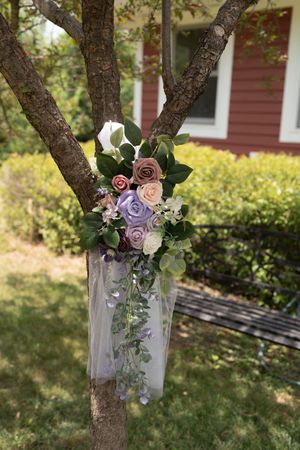 Elegant wedding floral arrangement of purple, pink, and white roses with greenery tied to a tree with white tulle.