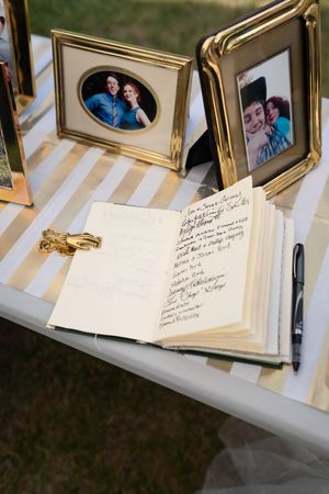 Wedding guest book open on a striped table with gold-framed couple photos and a pen at an outdoor ceremony.