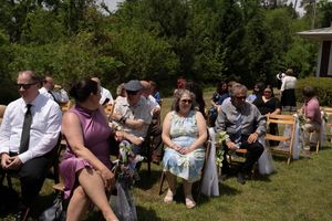 Wedding guests seated outdoors on chairs in sunny garden during wedding ceremony.