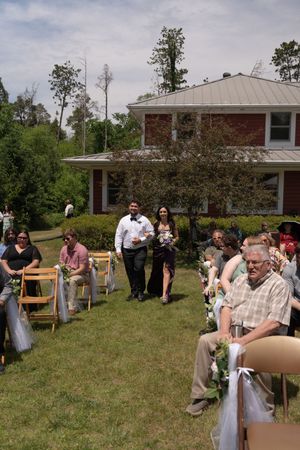 Wedding party processional walks down outdoor aisle between seated guests at backyard ceremony.