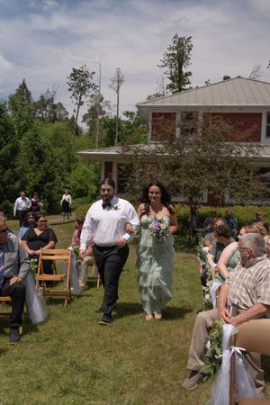 Bridesmaid in sage green dress walking down outdoor aisle with groomsman at garden wedding.
