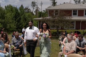 Bridesmaid in green gown walks down outdoor wedding aisle with groomsman past seated guests on sunny day.