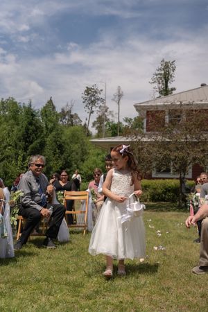 Young flower girl in white dress scattering petals down aisle at sunny outdoor wedding.