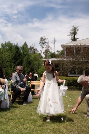 Young flower girl in white dress scatters petals during outdoor wedding ceremony on sunny day.