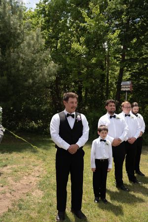 Groom and groomsmen in black vests and bow ties stand outdoors awaiting wedding ceremony to begin.