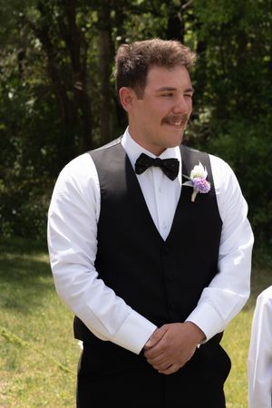 Smiling groom in black vest and bow tie with boutonniere enjoys a candid moment during outdoor wedding ceremony.