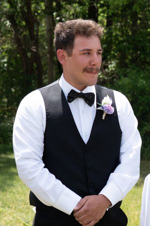 Handsome groom with mustache wearing black vest, bow tie, and floral boutonniere smiles outdoors at wedding.