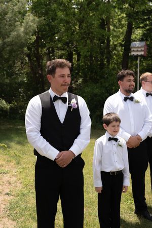 Groom in black vest and bow tie stands with groomsmen and ring bearer at outdoor wedding ceremony.