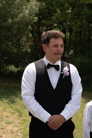 Emotional groom in black vest and bow tie with boutonniere awaits bride at outdoor ceremony.