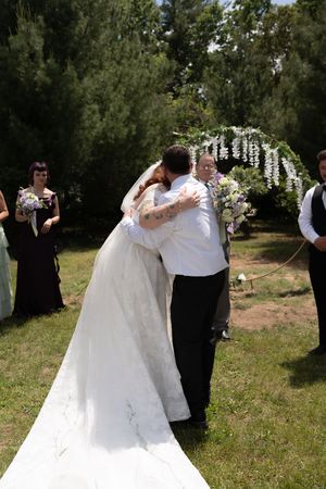 Bride and groom share emotional embrace at outdoor wedding ceremony with bridesmaids in black dresses.