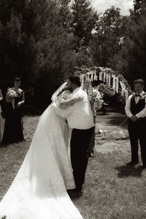 Black and white photo of bride and groom sharing first kiss at outdoor wedding ceremony under floral arch.