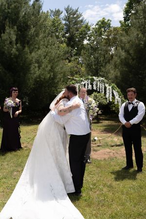 Bride and groom share first kiss at outdoor altar with wedding party and floral arch backdrop.