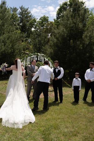 Bride in white gown walks toward groom and wedding party during outdoor garden ceremony.