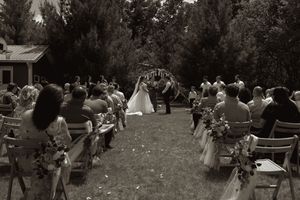 Black and white photo of outdoor wedding ceremony with bride walking down aisle toward floral arch and guests.