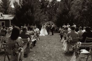 Black and white photo of an outdoor wedding ceremony with guests seated along a flower-lined aisle leading to the altar.