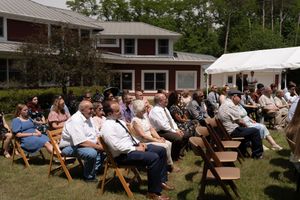 Guests seated in wooden folding chairs on a sunny lawn during an outdoor ceremony near a brick building.