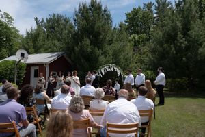 Outdoor wedding ceremony with guests seated on wooden chairs facing couple at a floral arch near a red barn.