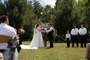 Outdoor wedding ceremony with bride and groom exchanging vows under circular floral arch surrounded by wedding party.