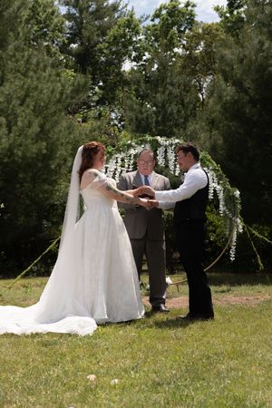 Bride and groom exchange vows outdoors under a floral arch with an officiant in a lush green garden setting.