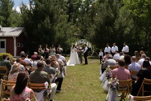 A bride and groom exchange vows under a floral arch at an outdoor wedding surrounded by seated guests and trees.