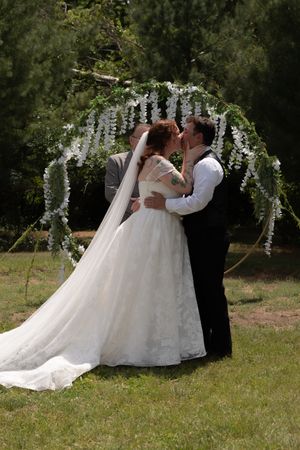 Bride and groom share their first kiss under a floral arch adorned with white wisteria at an outdoor wedding.
