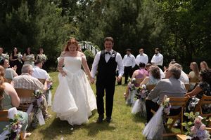Bride and groom walk hand-in-hand down the aisle at an outdoor wedding ceremony surrounded by guests.