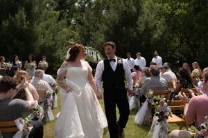 Newlyweds walk down the outdoor wedding aisle surrounded by seated guests in a garden setting.