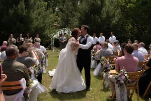 Bride and groom share first kiss at outdoor wedding ceremony with guests seated in rows on sunny day.