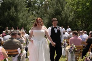 Newlywed couple walks down outdoor wedding aisle, bride in white gown and groom in black tuxedo, guests seated among trees.