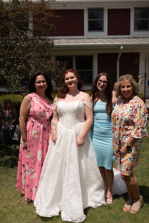 Four women posing outdoors, one in a white wedding gown, others in pink, blue, and floral dresses.