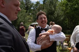 Groom in black vest and bow tie laughing while holding a baby at an outdoor wedding reception.