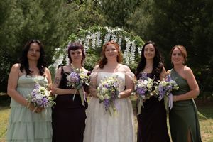Bride in white gown with four bridesmaids holding floral bouquets outdoors near a floral arch.