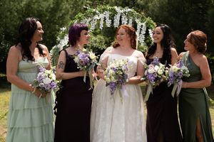 Bride with four bridesmaids in green dresses holding purple and white bouquets under a floral wedding arch.