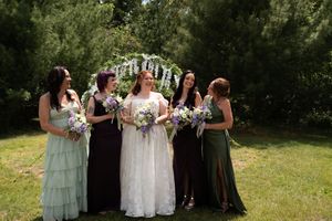 Bride with bridesmaids in varied green and purple gowns holding bouquets under floral arch outdoors.