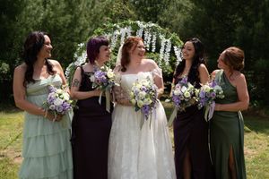 Bride in white lace gown stands with four bridesmaids in purple and green dresses holding bouquets under floral arch.