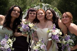 Bride and four bridesmaids in mismatched dresses smile together holding purple and white bouquets before a floral arch.