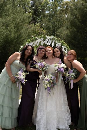 A bride in a white lace gown poses with four bridesmaids in green and purple dresses under a floral arch.