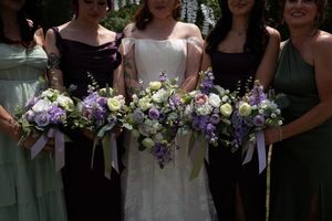 Bride and bridesmaids holding elegant purple and white floral bouquets with ribbon accents at an outdoor wedding.