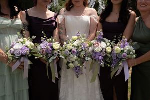Bride and bridesmaids in mismatched green and purple dresses display elegant lavender and white floral bouquets outdoors.