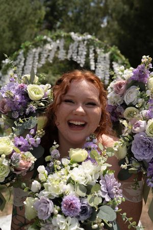 Joyful red-haired bride laughing surrounded by purple and white wedding bouquets under floral arch.
