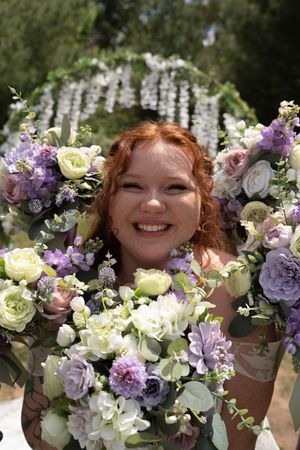 Smiling red-haired bride surrounded by lush purple and white floral bouquets at an outdoor wedding.