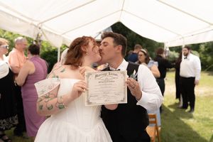 Bride and groom kissing while holding marriage certificate under white tent surrounded by wedding guests.